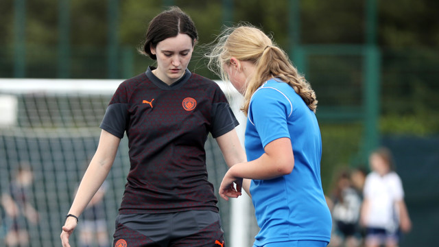 Two women soccer players on the field, one in a black kit and the other in a blue kit, standing close together with a net in the background.