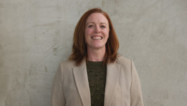 A woman wearing a beige blazer over a green knitted top, standing against a textured wall with a blurred face.