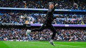 Person in a black tracksuit jumping in celebration on a football field at Etihad Stadium with a crowd in the background.