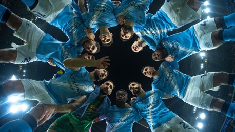 A group of football players in light blue jerseys and white shorts forming a huddle on the pitch.