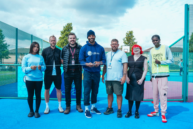 Group of seven people standing on a blue sports court, participating in a ribbon cutting ceremony. They are holding scissors and a ribbon, behind a fenced sports area.
