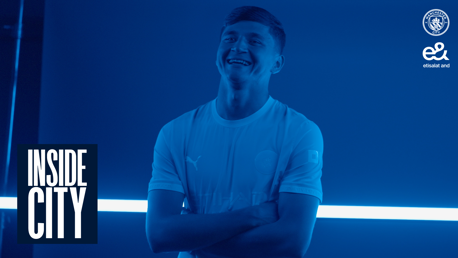 A person in a Manchester City shirt stands smiling with arms crossed in a blue-tinted image. The Manchester City logo and 'Inside City' text are visible.