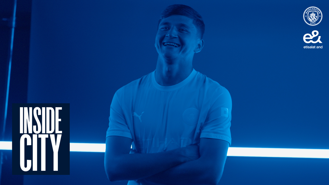 A person in a Manchester City shirt stands smiling with arms crossed in a blue-tinted image. The Manchester City logo and 'Inside City' text are visible.