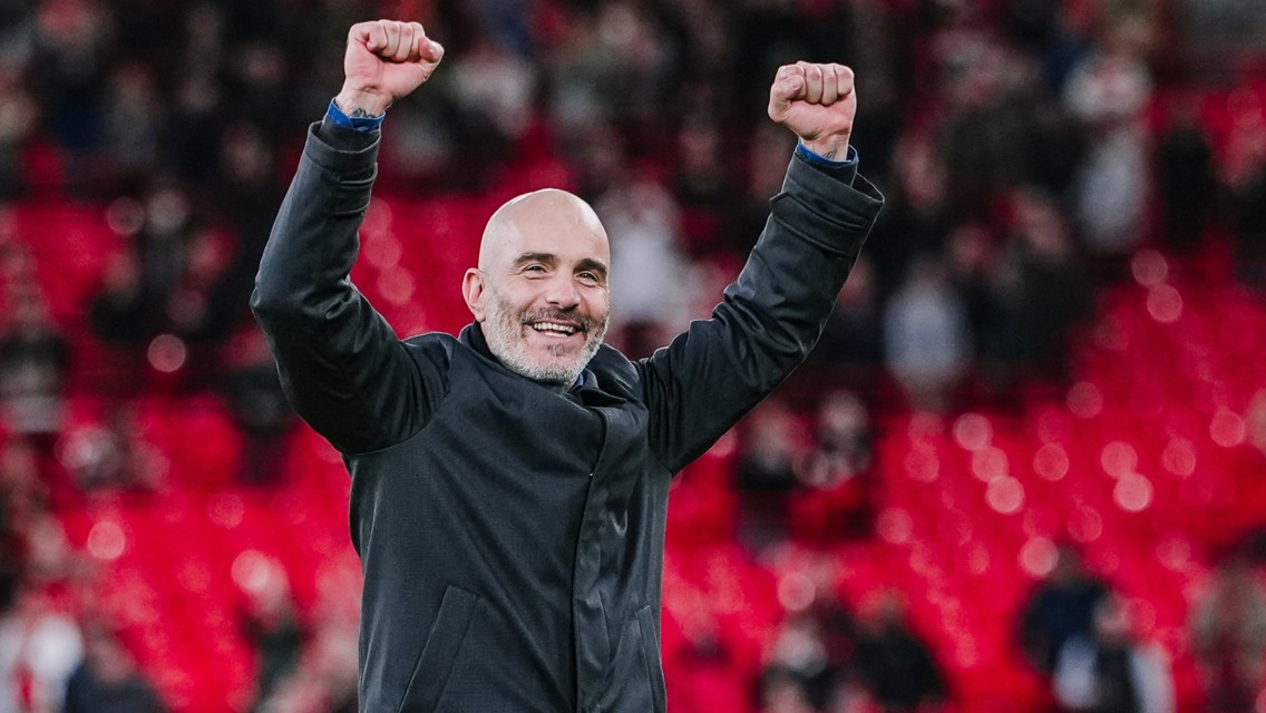 A man in a black jacket celebrating with raised fists in a stadium.