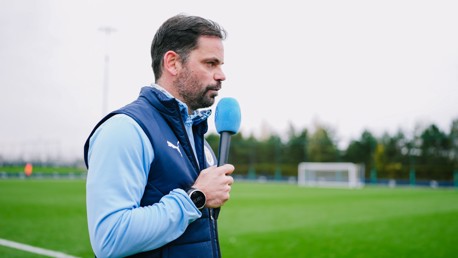 A person in a blue vest is holding a microphone on a soccer field. The field has goal posts and trees in the background.