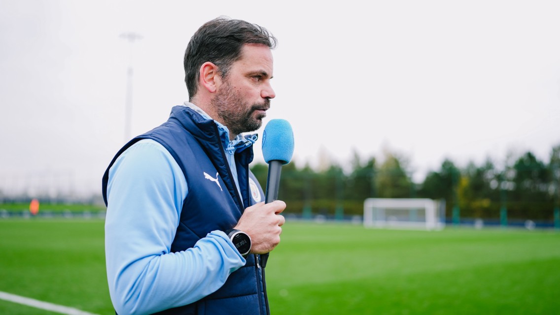 A person in a blue vest is holding a microphone on a soccer field. The field has goal posts and trees in the background.