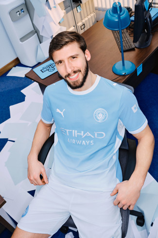 A person wearing the 2023 Manchester City home kit, seated in an office setting with papers scattered around.