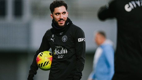 A Manchester City football player in black training attire holding a yellow ball with Manchester City logo visible on attire. Blurred faces of two individuals in the image.