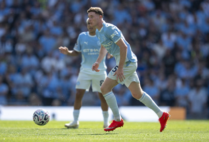 Two players in light blue Manchester City kits are on the field during a match. One is in motion with the ball nearby.