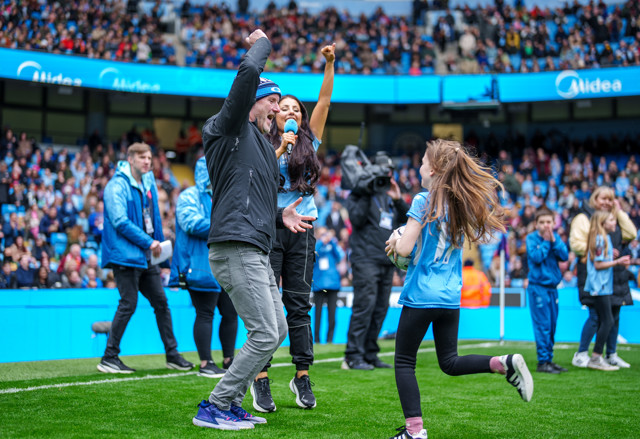 A man and a girl in Manchester City attire celebrate on a grassy field amidst a crowd at a stadium. The man raises his arm in triumph while the girl runs with a ball. Other people and a cameraman are visible in the background.