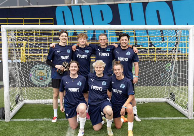 A soccer team poses in front of a goal, wearing blue jerseys with 'FOUDYS' and 'PUMA' branding. The stadium has a Manchester City crest in the background.