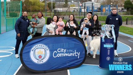 Group of people posing with Manchester City banner and Carabao Cup 2026 trophy outdoors on a sports court.