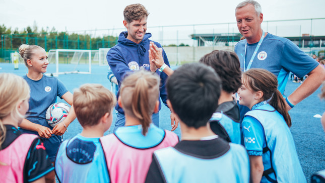 A group of children in football kits are engaged in a coaching session on a blue sports field. Two coaches wearing Manchester City apparel are animatedly interacting with the kids, providing guidance and encouragement.