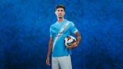 A person wearing a Manchester City jersey holding a football against a blue textured background.