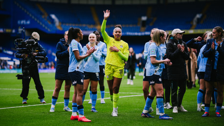A group of Manchester City Women players on the field, wearing team kits. One player in the center waves while others clap.