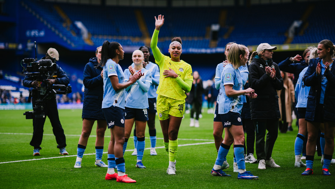 A group of Manchester City Women players on the field, wearing team kits. One player in the center waves while others clap.