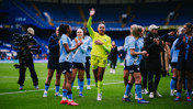 A group of Manchester City Women players on the field, wearing team kits. One player in the center waves while others clap.