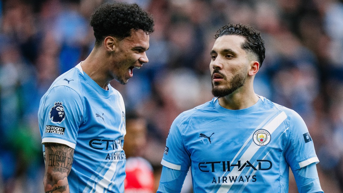 Two Manchester City players in conversation on the field wearing the team's sky blue uniform with the Etihad Airways sponsor logo.