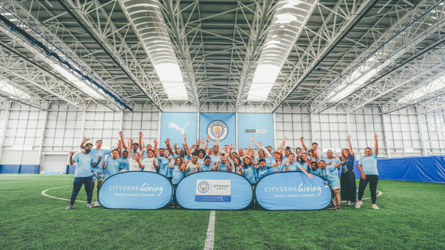 A diverse group of young adults in light blue shirts celebrate at a sporting event inside a Manchester City training facility, showcasing banners for Cityzens Giving and Etihad Airways.