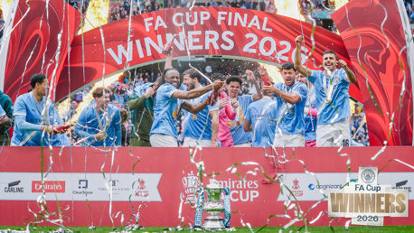 Manchester City players celebrating FA Cup victory in front of a banner with fireworks and confetti, showing the trophy at Wembley Stadium.