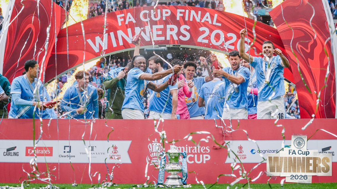 Manchester City players celebrating FA Cup victory in front of a banner with fireworks and confetti, showing the trophy at Wembley Stadium.