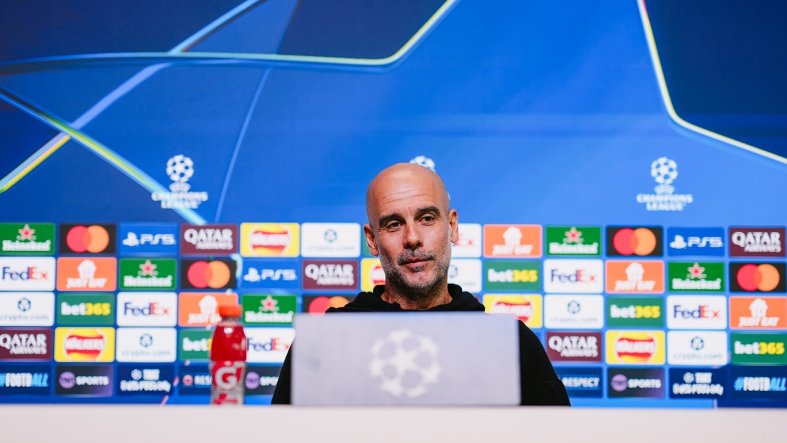 A blurred face is at a press conference setup with UEFA Champions League branding and sponsor logos. A Gatorade bottle is visible in the foreground.