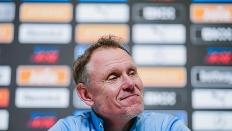 A person with a blurred face sitting at a press conference table. Behind them is a backdrop featuring various logos and sponsors.