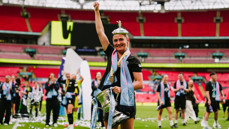 Player holding a trophy in a football stadium with teammates celebrating in the background.