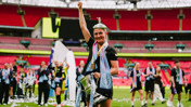 Player holding a trophy in a football stadium with teammates celebrating in the background.