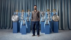 A man standing among various football trophies including multiple league cups, Premier League trophies, and the FA Community Shields.