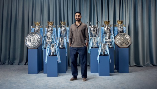 A man standing among various football trophies including multiple league cups, Premier League trophies, and the FA Community Shields.