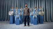 A man standing among various football trophies including multiple league cups, Premier League trophies, and the FA Community Shields.
