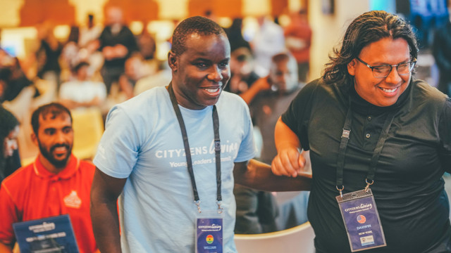 Two individuals at a CityzensGiving Young Leaders Summit event, wearing badges with names and country flags, engaged in a conversation or activity. Other attendees are visible in the blurred background.