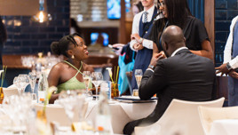 A group of people seated at a restaurant table, smiling and conversing with a waiter in formal attire standing nearby.