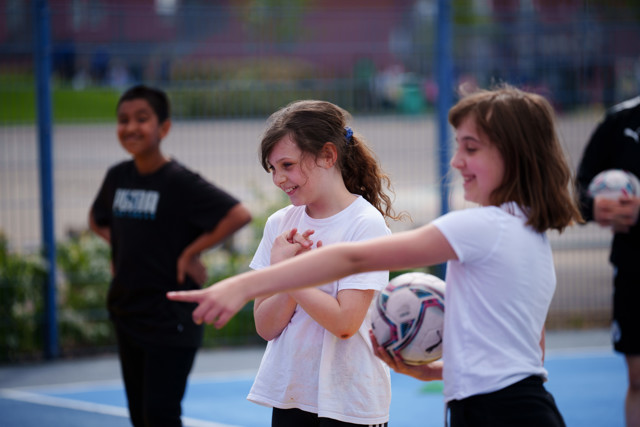 A group of children wearing athletic attire interacting with a ball outdoors in a fenced area. One child is pointing while another holds the ball.
