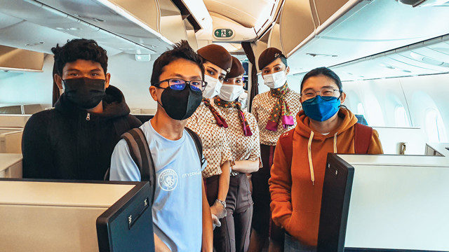 A group of people including two flight attendants in uniform, standing inside an aircraft cabin. Visible elements include seats and overhead compartments.