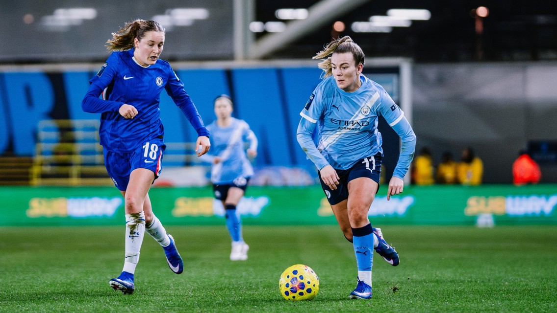 Football players in blue and light blue kits are in action, focusing on a yellow ball on the field.