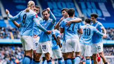 Manchester City players celebrating a goal at Etihad Stadium. Players are wearing light blue jerseys with names and numbers visible on the back.