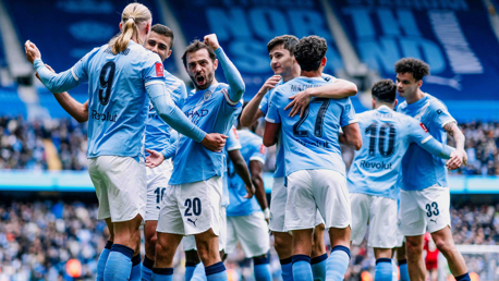 Manchester City players celebrating a goal at Etihad Stadium. Players are wearing light blue jerseys with names and numbers visible on the back.