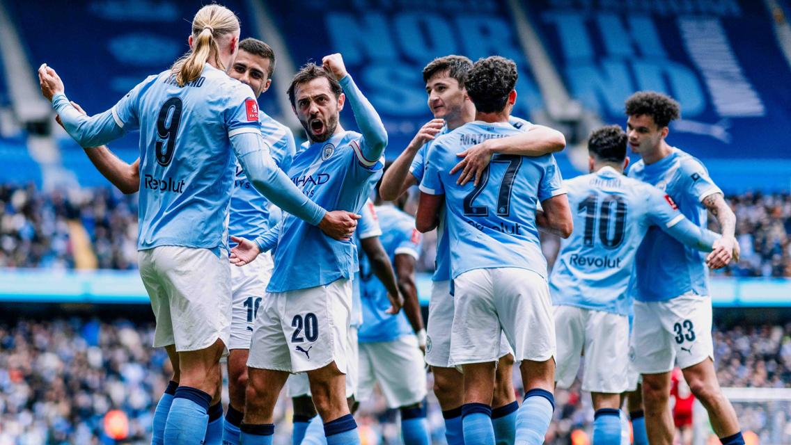 Manchester City players celebrating a goal at Etihad Stadium. Players are wearing light blue jerseys with names and numbers visible on the back.