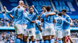 Manchester City players celebrating a goal at Etihad Stadium. Players are wearing light blue jerseys with names and numbers visible on the back.