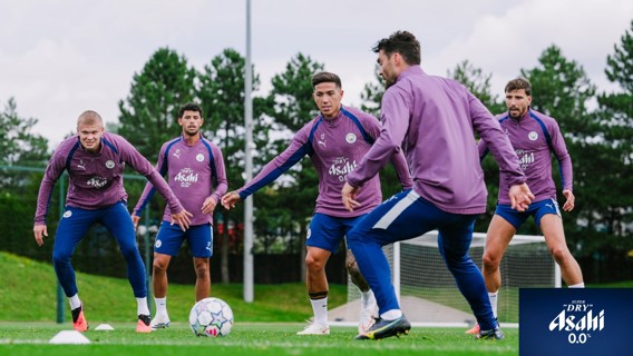Five football players in training attire with Manchester City logos practice on a field. They wear purple jackets with Asahi branding and engage in a training drill near goalposts.