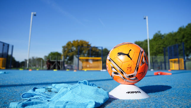 Orange Puma soccer ball placed on a white Kipsta marker cone on a blue training ground with blurred football nets and trees in the background under clear blue sky.