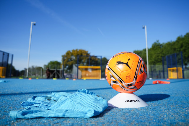 Orange Puma soccer ball placed on a white Kipsta marker cone on a blue training ground with blurred football nets and trees in the background under clear blue sky.