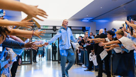 A player wearing a Manchester City tracksuit walks through a corridor, greeting fans who reach out excitedly.