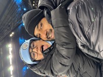 Two people dressed warmly in winter attire are attending a football match. One is wearing a Manchester City hat. The stadium lights are visible in the background.