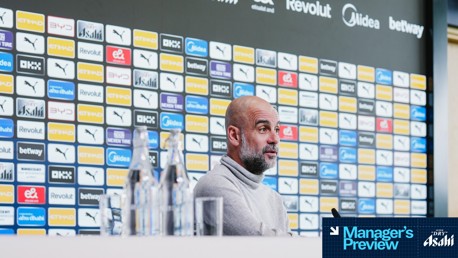 A person sits at a press conference table with blurred face. Background featuring various sponsor logos including Puma, Etihad, and Revolut. Water bottles arranged on the table. 'Manager's Preview' text with Asahi branding.