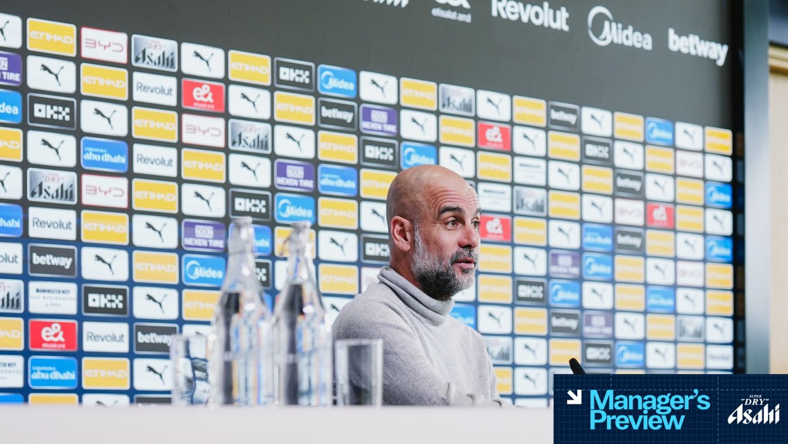 A person sits at a press conference table with blurred face. Background featuring various sponsor logos including Puma, Etihad, and Revolut. Water bottles arranged on the table. 'Manager's Preview' text with Asahi branding.