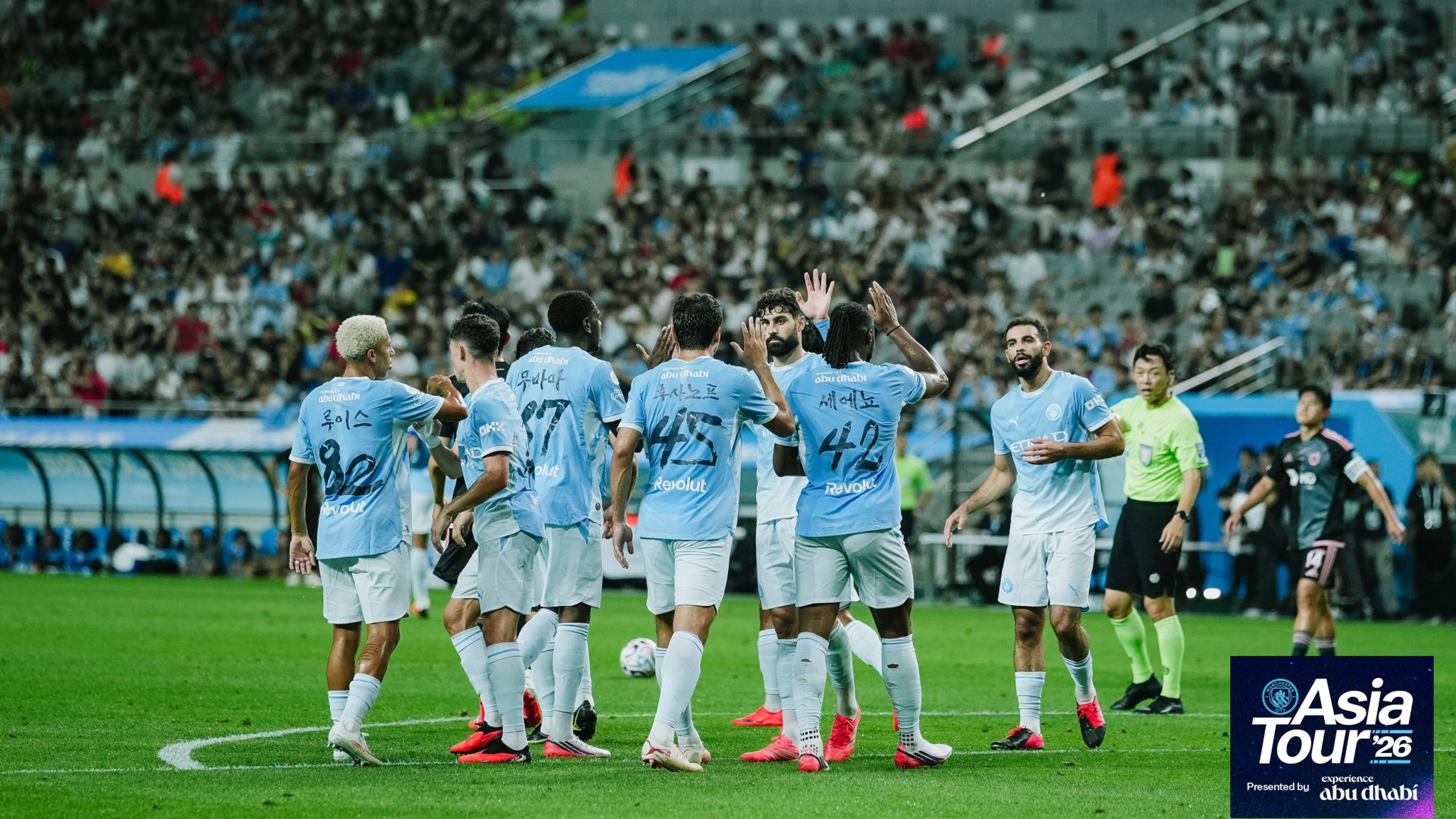 Manchester City players celebrating a goal during the Asia Tour '26, with branding for experience Abu Dhabi. The team is wearing light blue jerseys with logos and player names displayed in various languages.