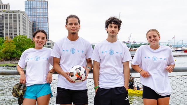 Four individuals wearing white t-shirts with logos, standing by a waterfront. The second person is holding a soccer ball.